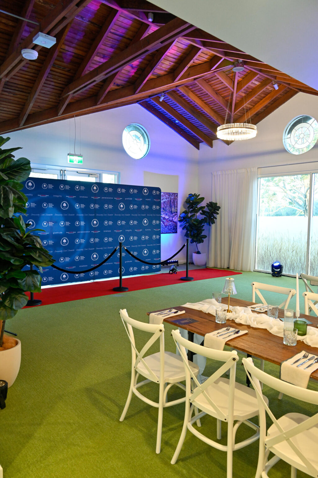 A room with a high wooden ceiling features a green carpet, white chairs around a wooden table, and a blue step-and-repeat banner with logos. There are potted plants and circular windows, with natural light coming in.