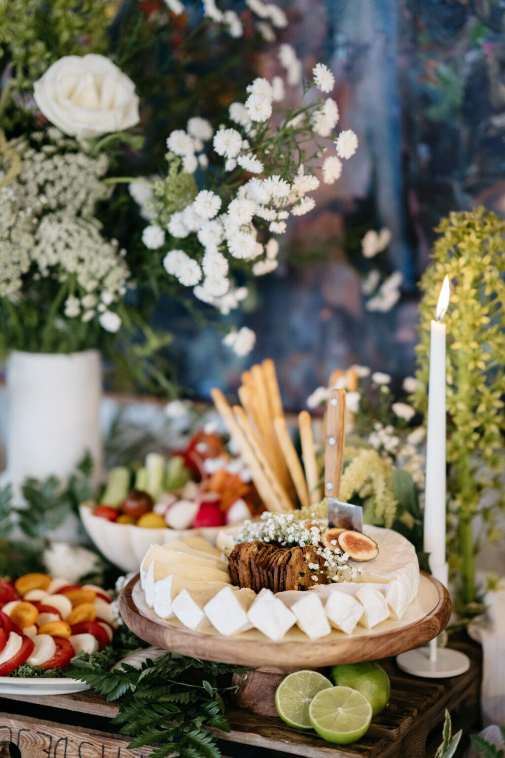 A cheese platter with brie, figs, crackers, and breadsticks on a wooden stand, surrounded by sliced fruit, limes, a lit candle, and a vase of white flowers and greenery in the background.