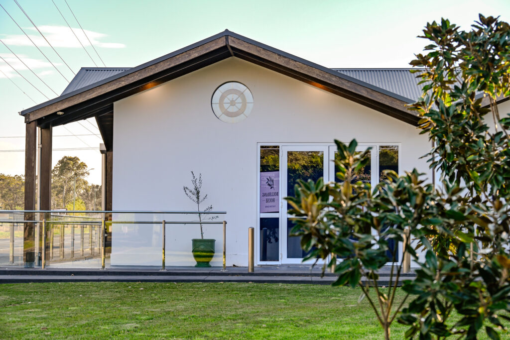 A modern single-story building with a white exterior, large glass doors, a small potted plant, and a decorative round feature above the entrance. Trees and grass are visible in the foreground and background.