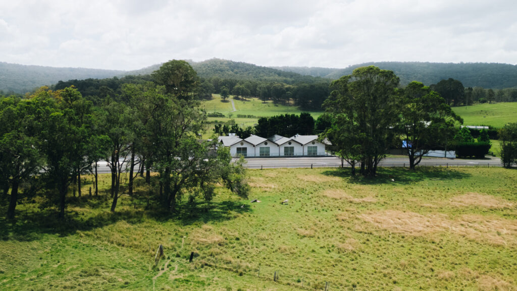 A row of white buildings is surrounded by trees and fields, with a road in front and hills covered in greenery in the background under a cloudy sky.