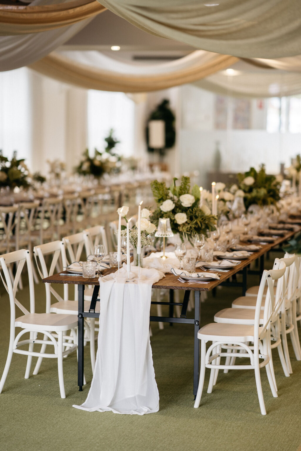 Elegant wedding reception table with white chairs, draped white fabric table runner, tall candles, lush white and green floral centerpieces, glassware, and neutral decor under beige fabric-draped ceiling.
