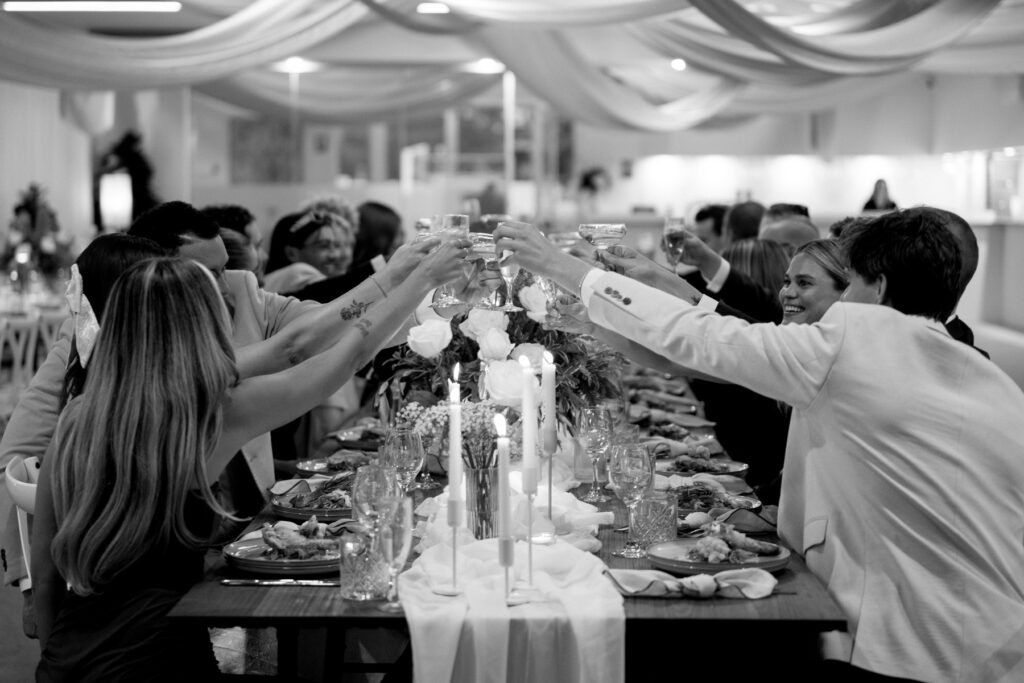 A group of people sit around a long, elegantly decorated table, raising their glasses in a toast. The setting is festive, with candles, flowers, and draped fabric overhead. The photo is in black and white.