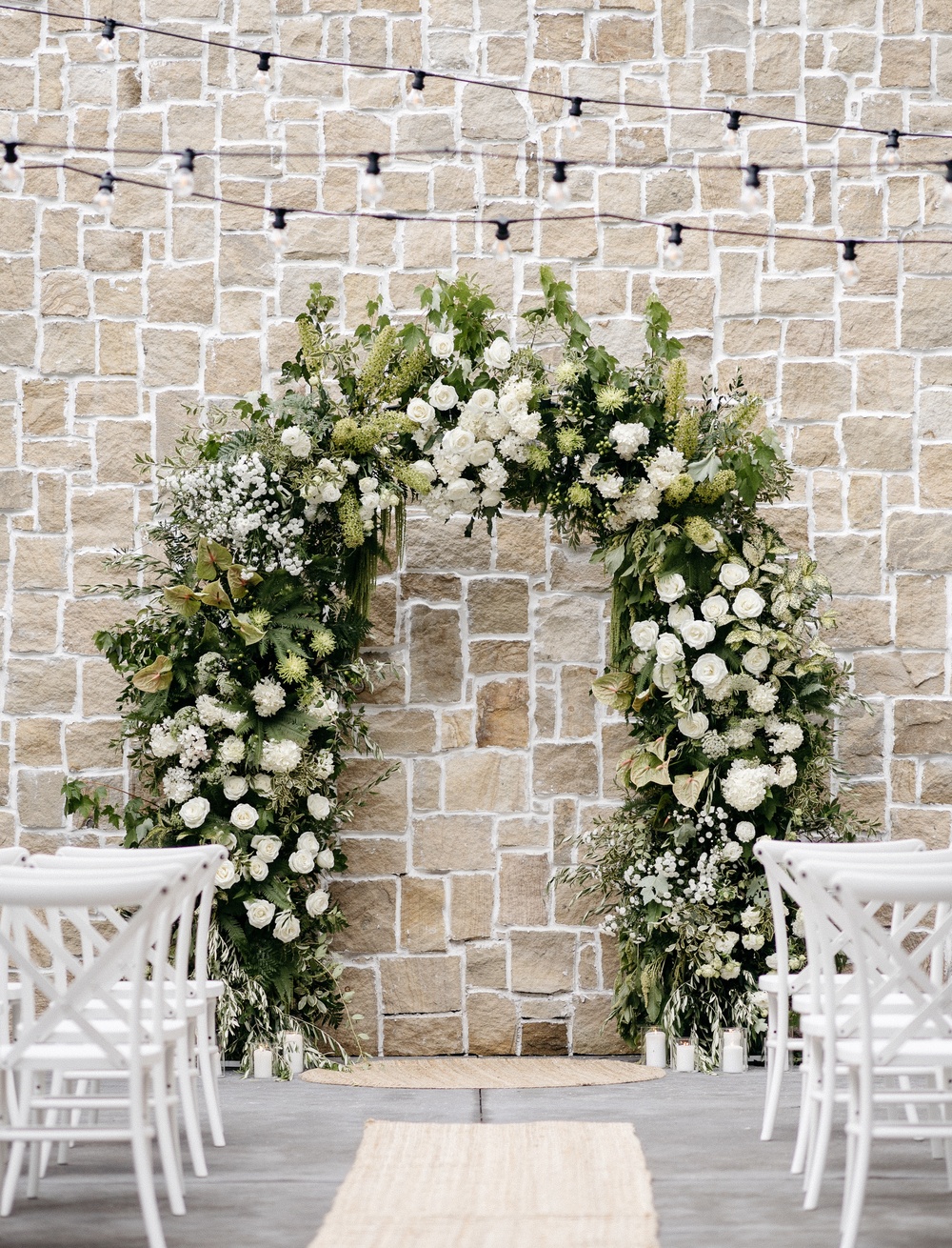 A floral arch adorned with white and green flowers stands in front of a stone wall at Bollerang House, a stunning South Coast wedding venue, with white chairs lining an aisle set with a beige runner and glowing string lights above.