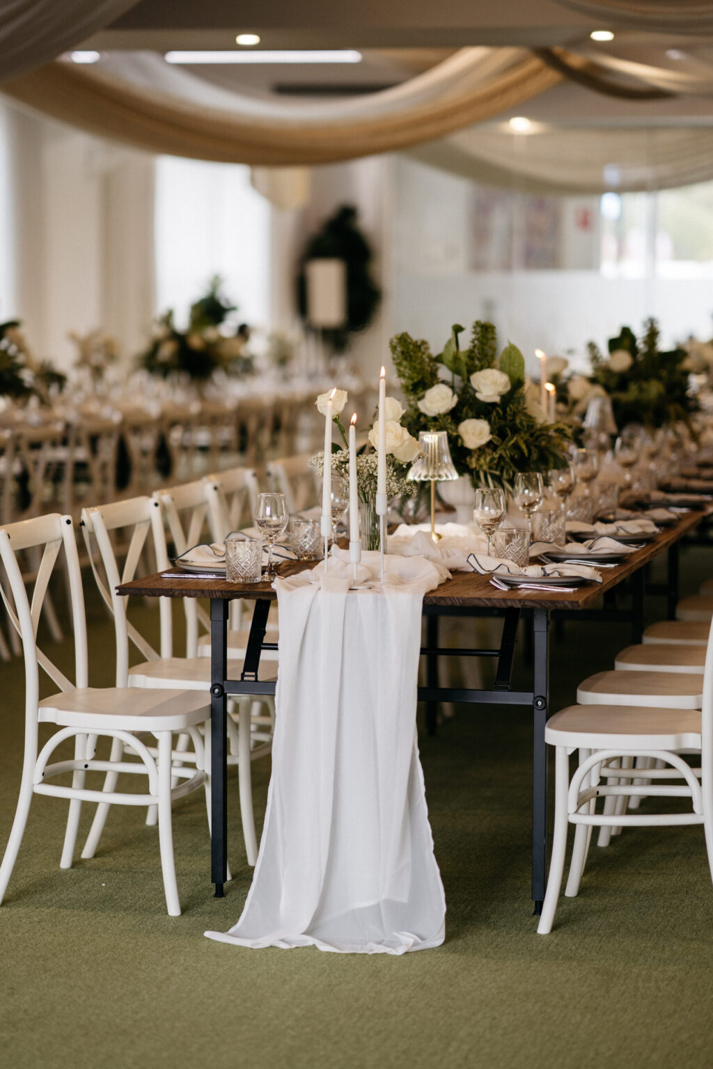 A long, elegant dining table is decorated with white cloth, tall candles, lush green and white floral centerpieces, and neatly arranged plates and glasses, set in a bright, decorated event space.