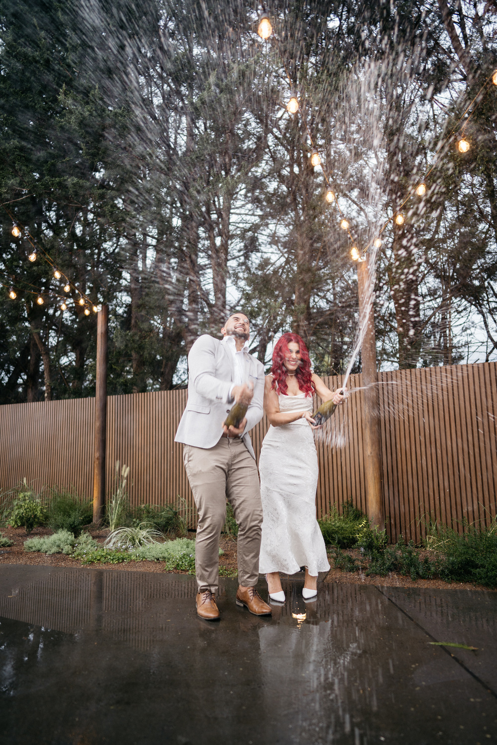 A couple dressed in formal attire joyfully pops a champagne bottle, with bubbly spray shooting into the air. They stand outside on a wet pavement, surrounded by trees and string lights.