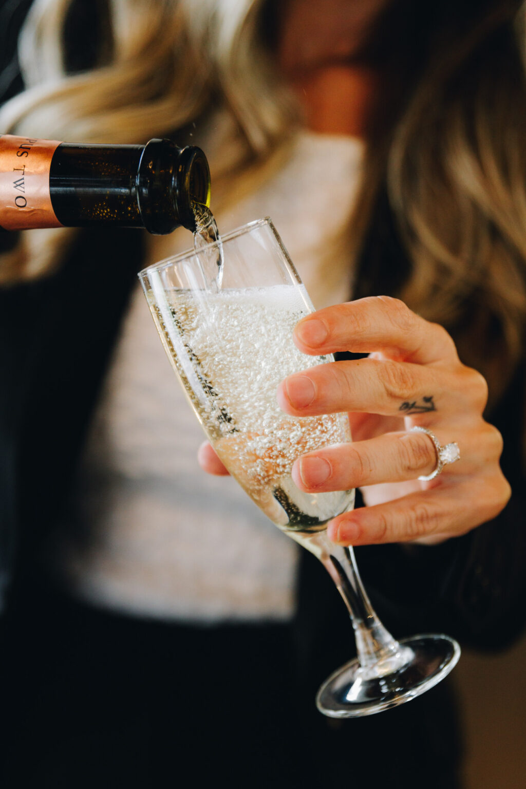 A person with long hair pours sparkling wine into a champagne flute, holding the glass with a hand that has a ring on the ring finger.