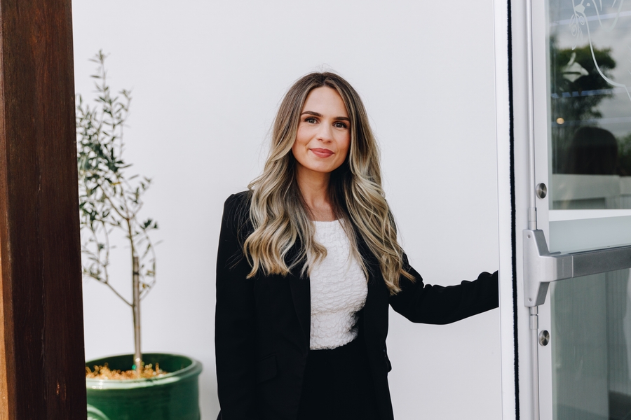A woman with long, wavy blonde hair wearing a black blazer and white top stands near an open glass door, smiling. There is a green potted plant and a white wall in the background.
