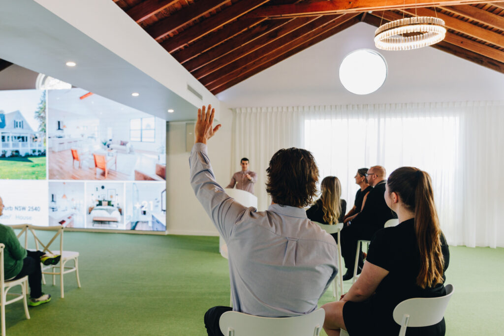 A man raises his hand while seated in an audience, listening to a speaker presenting in front of a large screen with images of houses, inside a modern, well-lit room with wooden ceiling beams.