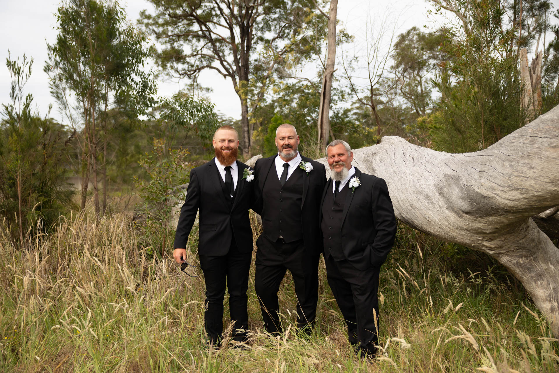 Three men in black suits and ties, each with a white buttonhole, stand together smiling in a grassy, wooded outdoor setting near a fallen tree at the Renae and Wade wedding. The logo “H. PARSONS” appears in the corner.