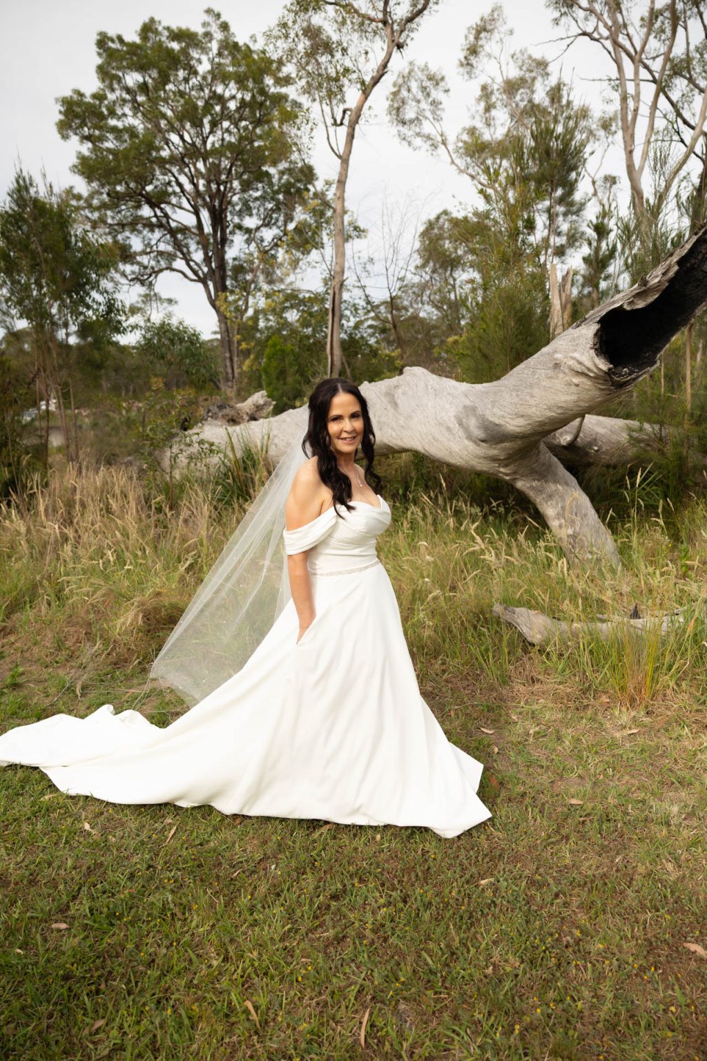 A woman in an off-the-shoulder white wedding dress and veil stands on grass in front of a large fallen tree and tall greenery, smiling at the camera.