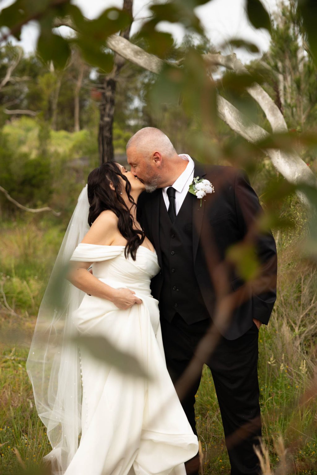A bride in an off-the-shoulder white dress and veil kisses a groom in a black suit and tie outdoors, framed by greenery and trees in the background.
