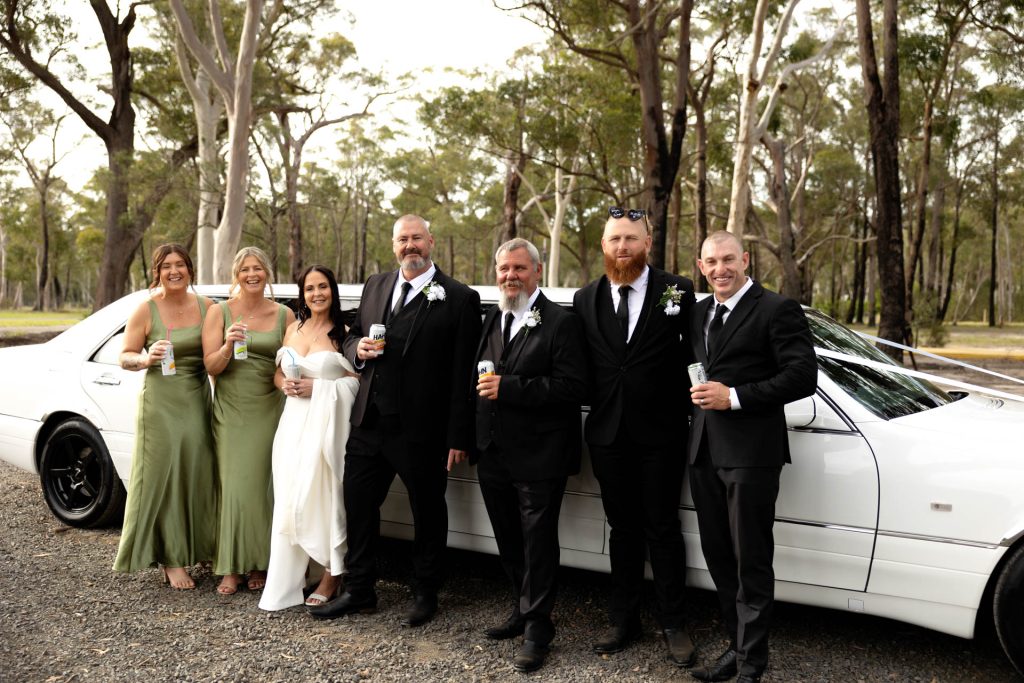 Six people in formal attire, three women and three men, stand in front of a white limousine in a wooded outdoor setting, smiling and holding drinks. The women wear green dresses and the men are in black suits.