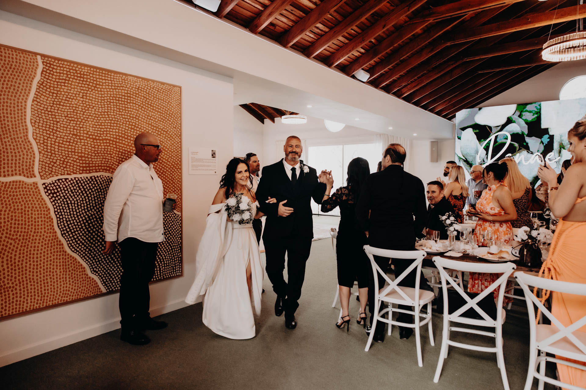 A smiling couple, arm in arm, walk into a bright reception room filled with seated guests. The woman wears a white dress with black details, and the man is in a dark suit—guests watch and smile at Renae and Wade's wedding celebration.