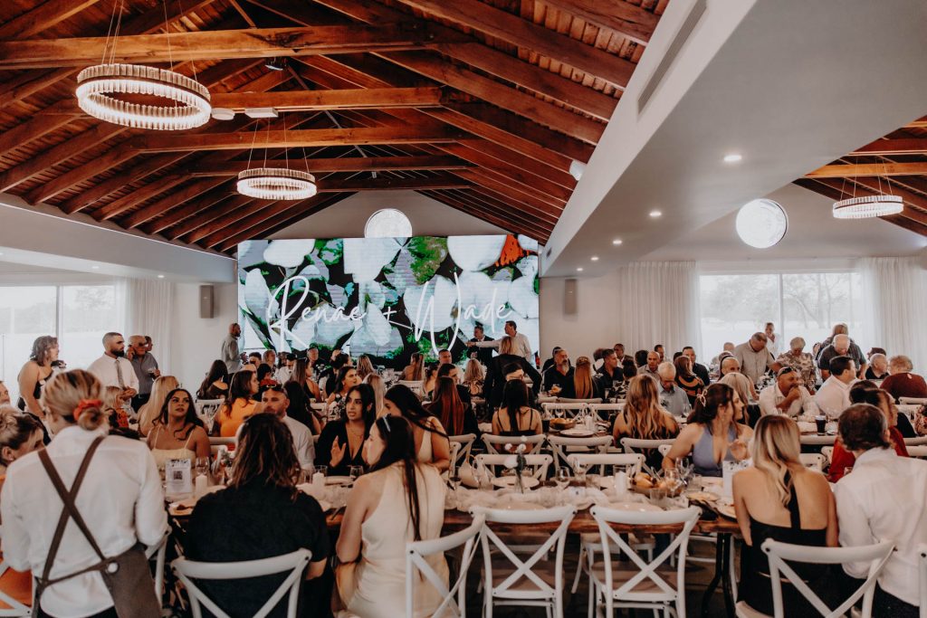 A large group of people are seated at tables in a modern event hall with a wooden ceiling. A screen at the front displays “Perfectly Paired.” The atmosphere is lively, with guests dining and socialising.