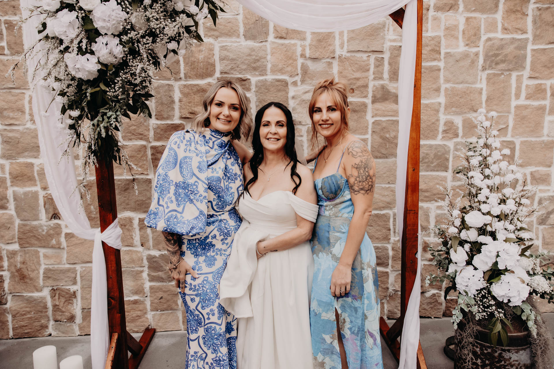 Three women in formal dresses stand smiling under a floral arch at the Renae and Wade wedding, in front of a stone wall. Floral arrangements and draped fabric decorate the scene. The logo "H. Parsons Funeral Directors" appears in the bottom right corner.