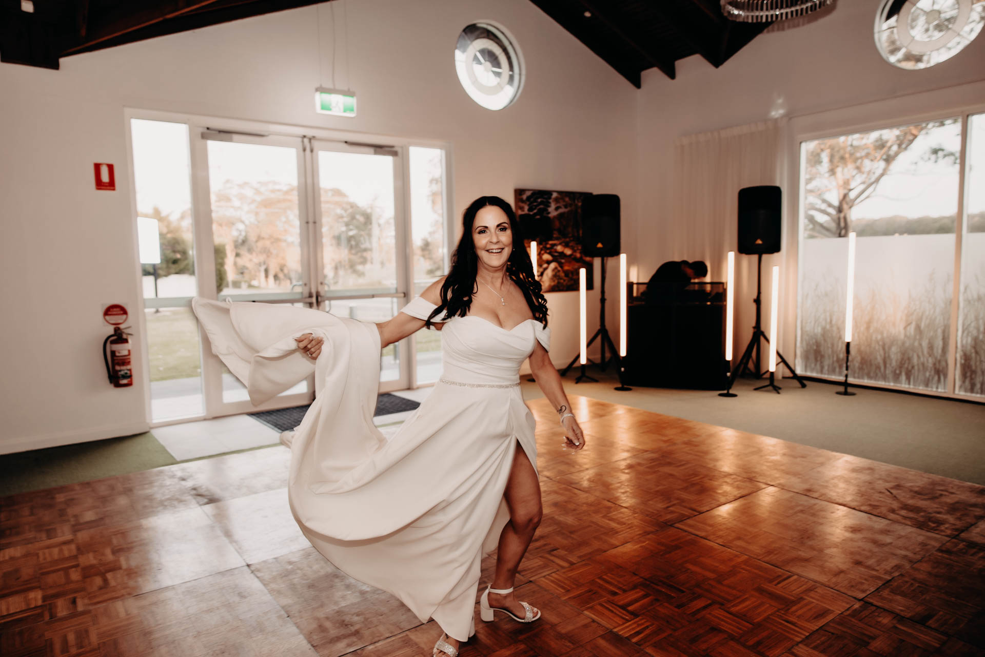 A smiling woman in a white, off-the-shoulder dress twirls on a wooden dance floor during the Renae and Wade wedding in a bright room with large windows. A fire extinguisher is on the wall and a "Funeral Directors H. Parsons" logo is in the corner.