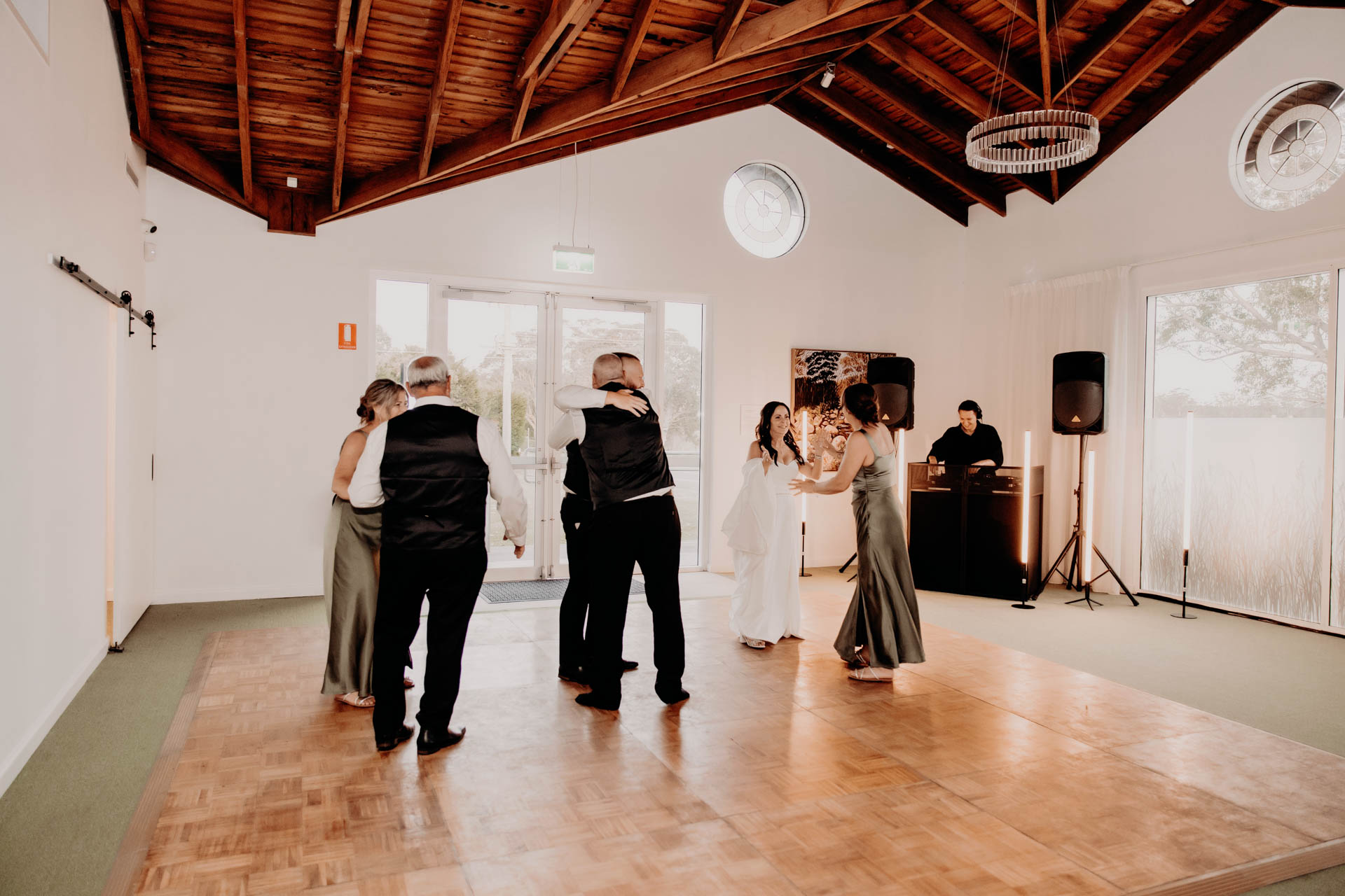 Three pairs of people, dressed in formal attire, dance on a wooden floor in a bright room at the Renae and Wade wedding, whilst a DJ stands at a table with speakers. The H. Parsons Funeral Directors logo is visible in the background.
