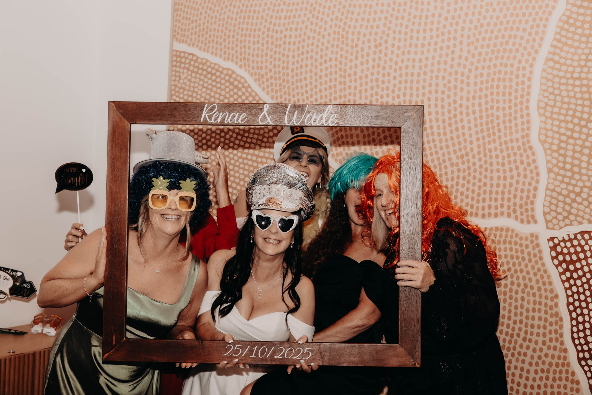 Five women in colourful costumes and novelty glasses smile whilst posing together, holding a large photo frame labelled “Renae & Wade, 25/10/2025,” capturing the fun spirit of the Renae and Wade wedding in front of a patterned wall.
