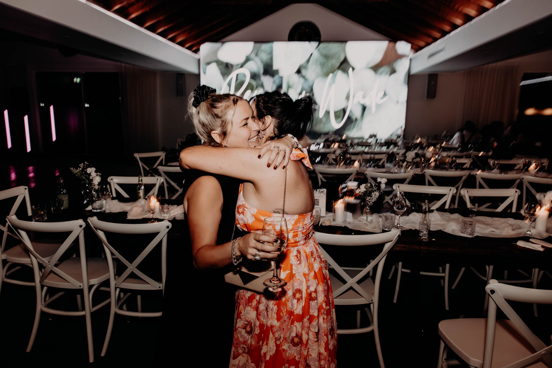 Two women hug and smile in a warmly lit event space with decorated tables. One holds a drink, whilst a projection in the background hints at the Renae and Wade wedding. The atmosphere is joyful and celebratory.