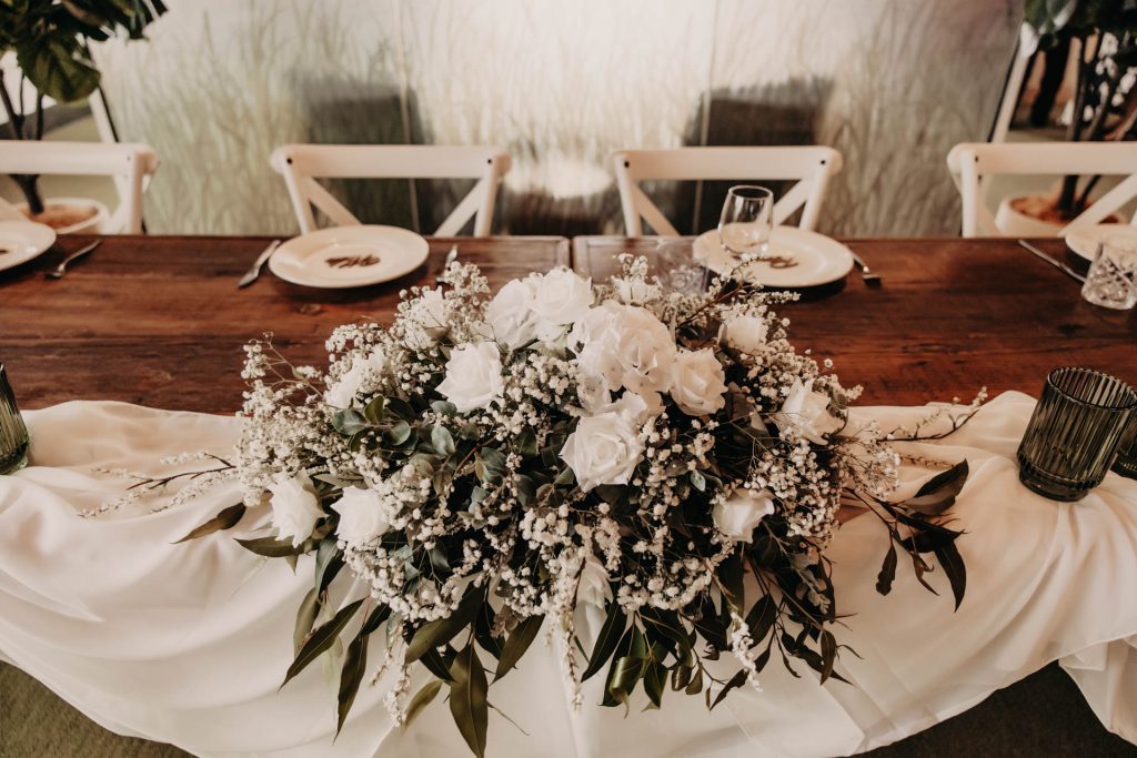 A wooden table decorated with a white floral centrepiece, featuring roses and greenery, atop a white cloth runner. Plates, glasses, and green glass tumblers are set at each seat, with white chairs in the background.