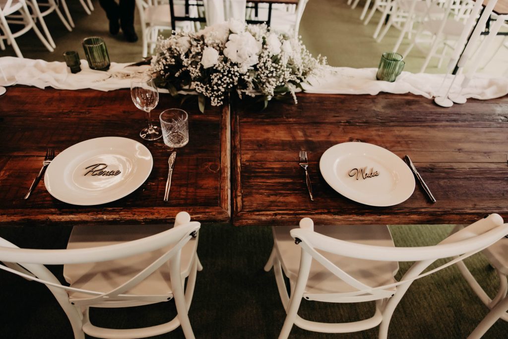 Two rustic wooden tables set side by side, each with a white chair and a plate labelled "Groom" and "Bride." White flowers and greenery decorate the centre, and place settings include glasses and cutlery.