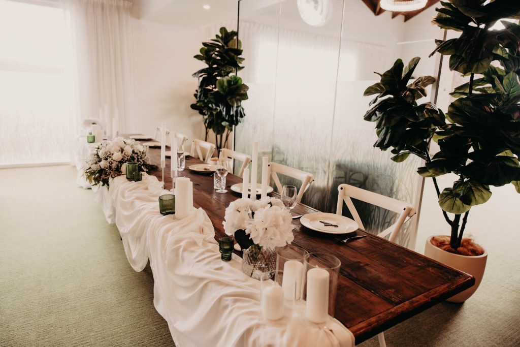 A long wooden table decorated with white fabric, candles, glass vases, and white flowers, set for a formal event in a bright room with green plants and white chairs.