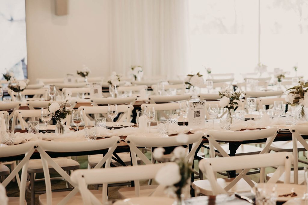 Elegant function room with rows of white chairs and tables set with glassware, plates, and white floral centrepieces. Bright natural light fills the airy, sophisticated space.