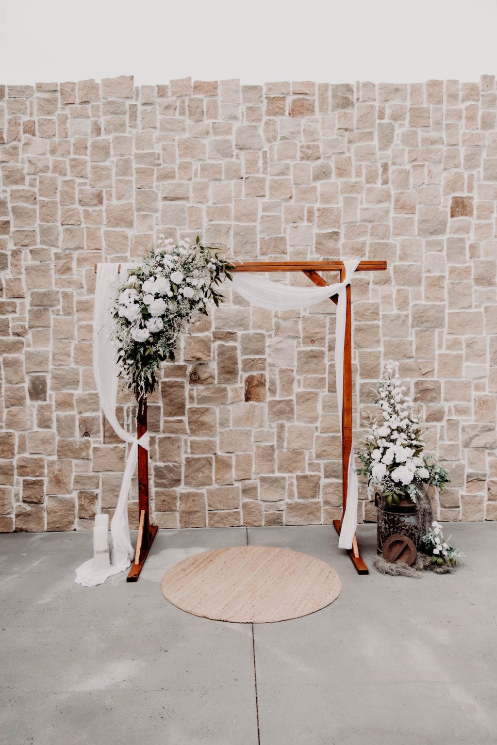 A wooden wedding arch decorated with white flowers and white fabric stands in front of a stone wall. A round beige rug is on the floor, and there are more white flowers and greenery in a basket to the right of the arch.