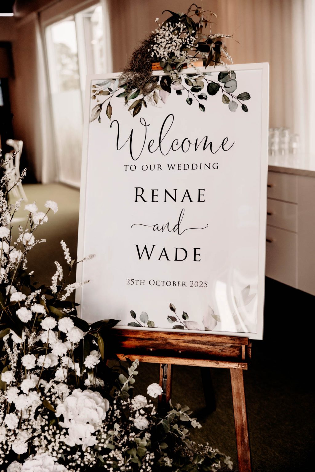 A white wedding welcome sign on an easel, decorated with greenery and flowers, reads: “Welcome to our wedding Renae and Wade, 25 October 2025.” White flowers are arranged around the base.