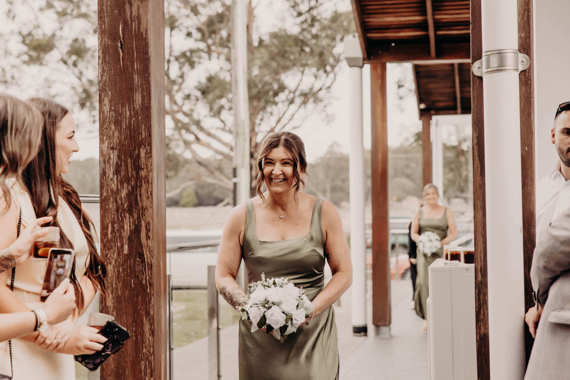 A smiling bridesmaid in a sage green dress walks down an outdoor aisle holding a white bouquet at the Renae and Wade wedding, whilst guests watch and take photos. The setting is bright, surrounded by trees. H. Parsons Funeral Directors logo is visible.
