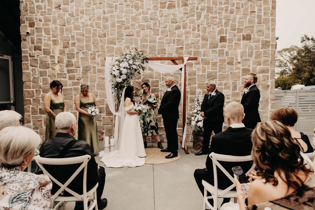 A bride and groom stand under a floral wedding arch during an outdoor ceremony, surrounded by bridesmaids in green dresses, groomsmen in dark suits, and seated guests facing them.