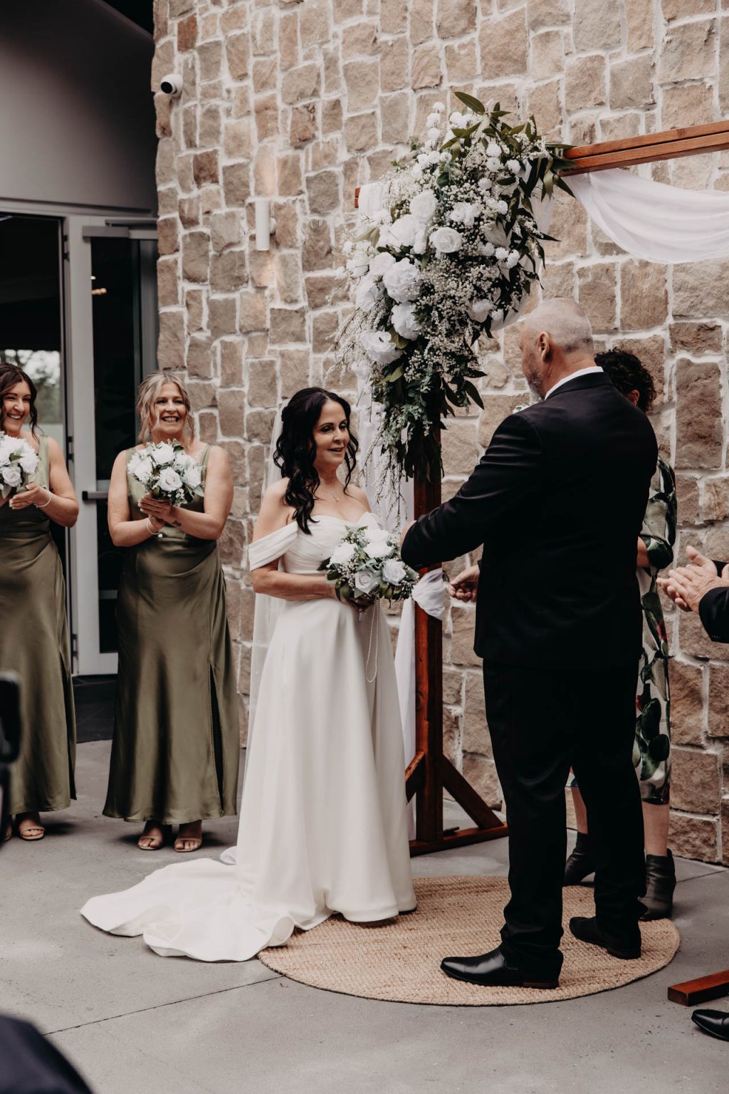 A bride and groom stand facing each other, holding hands during an outdoor wedding ceremony. Bridesmaids in green dresses and floral bouquets stand nearby in front of a stone wall and a white-and-green flower arrangement.
