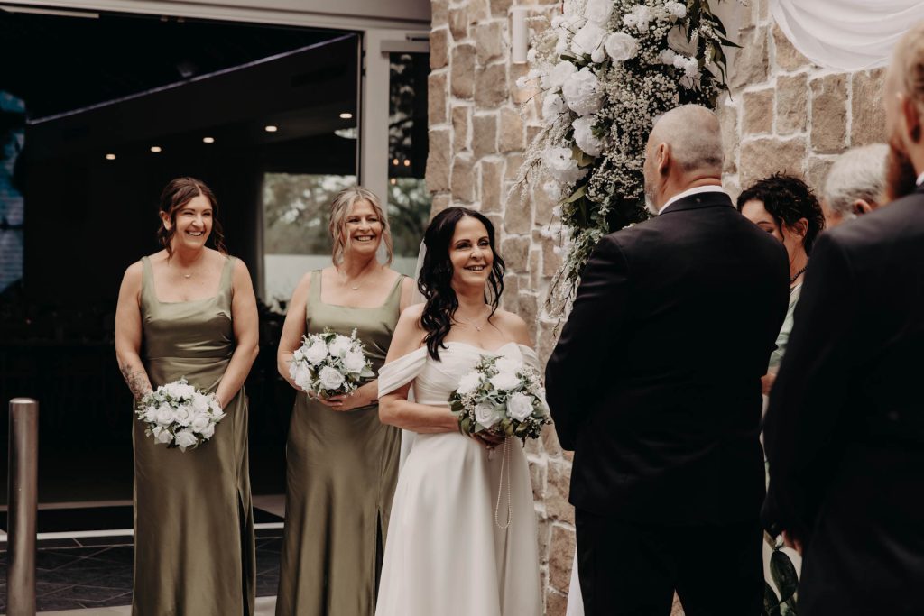 A bride in a white dress smiles at the groom during an outdoor wedding ceremony, with two bridesmaids in sage green dresses holding bouquets and standing beside her. White flowers decorate the stone wall behind them.