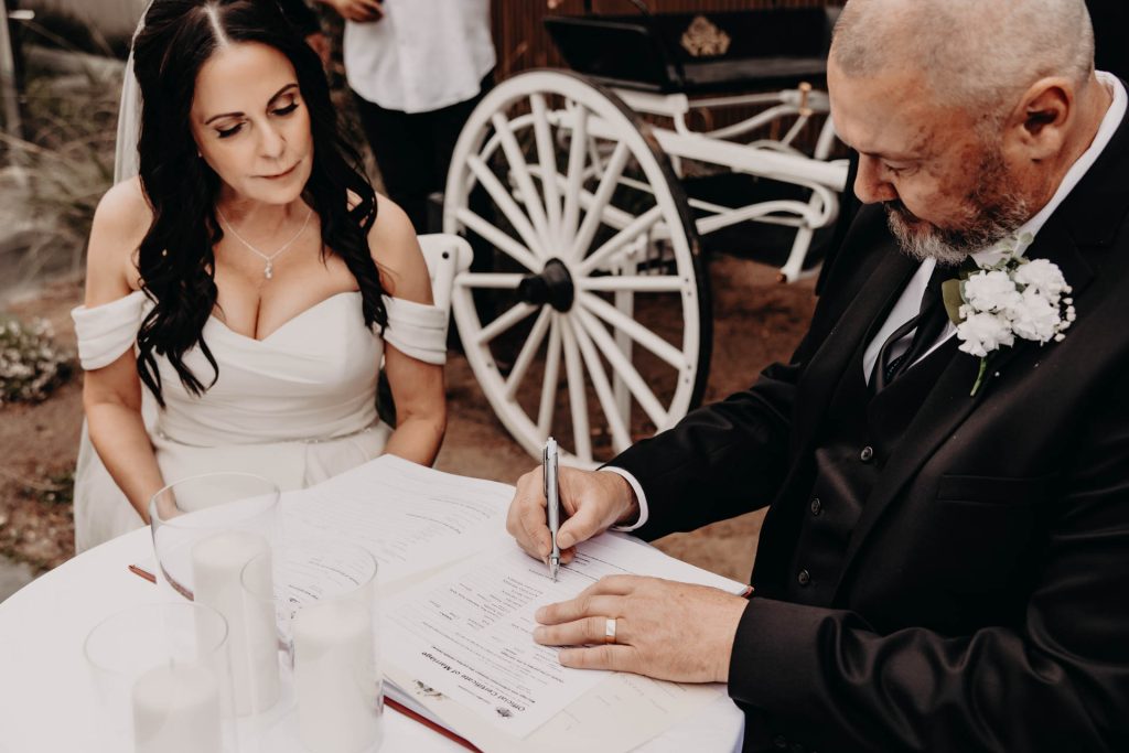 A man in a suit signs a document at a table while a woman in a white off-shoulder dress and veil watches. Three glass candles are on the table, and a white carriage wheel is visible in the background.