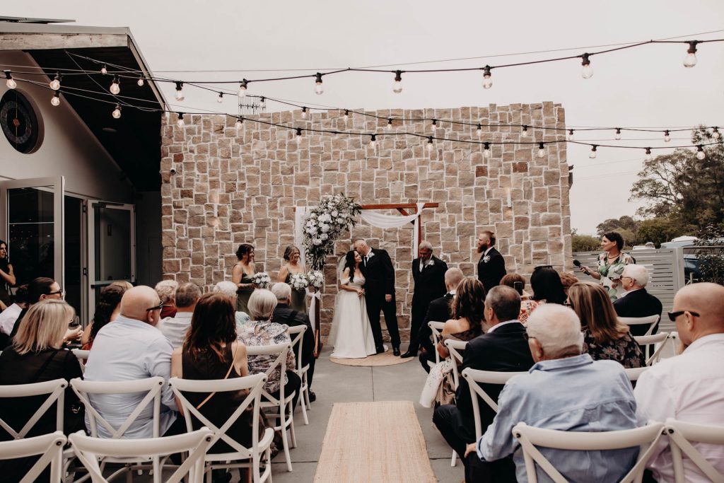 An outdoor wedding ceremony with a couple exchanging vows under a flower-decorated arch, surrounded by bridesmaids, groomsmen, and seated guests on white chairs, in front of a stone wall and fairy lights.