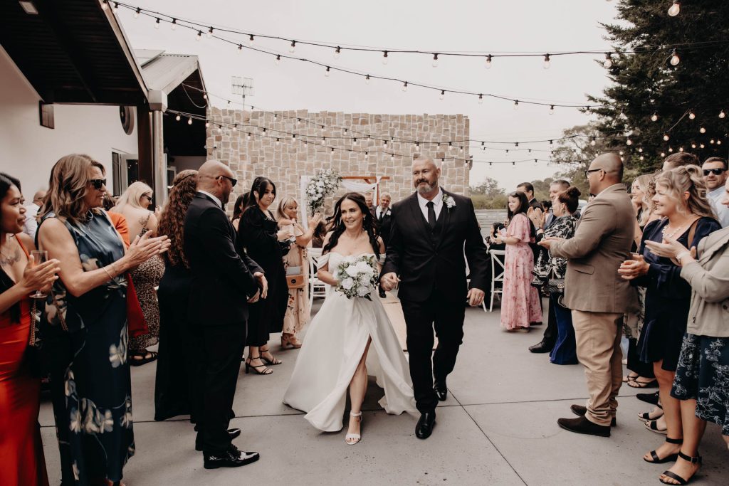 A bride in a white dress holding a bouquet walks arm-in-arm with a man in a dark suit down an outdoor aisle, surrounded by applauding guests under fairy lights.