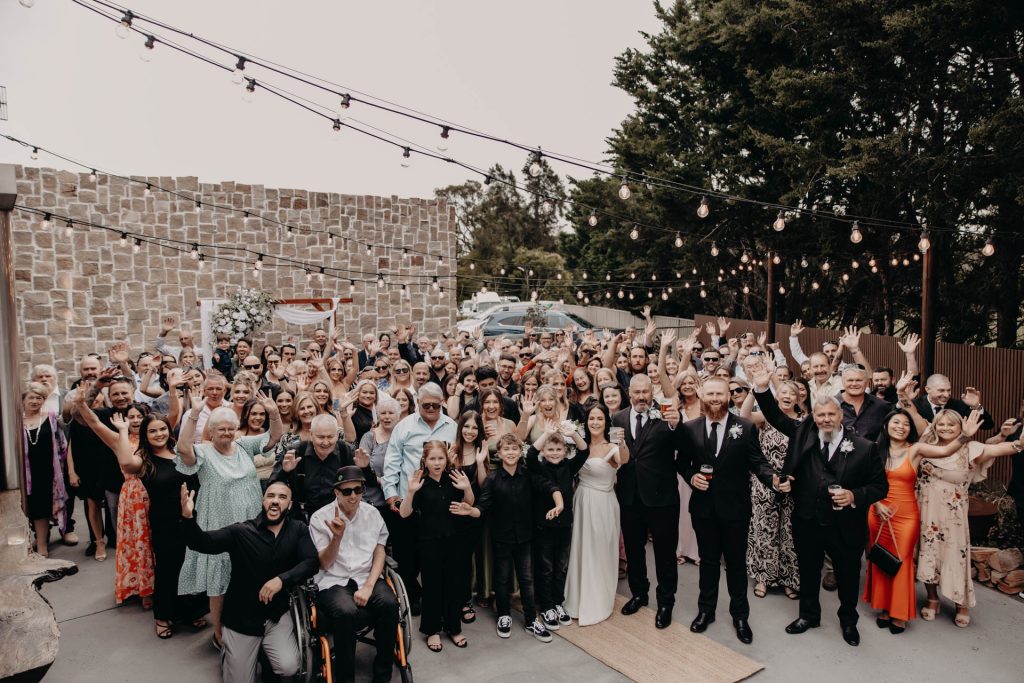 A large group of people, dressed in semi-formal attire, smile and wave at the camera at an outdoor event with fairy lights, a stone wall, and greenery in the background.