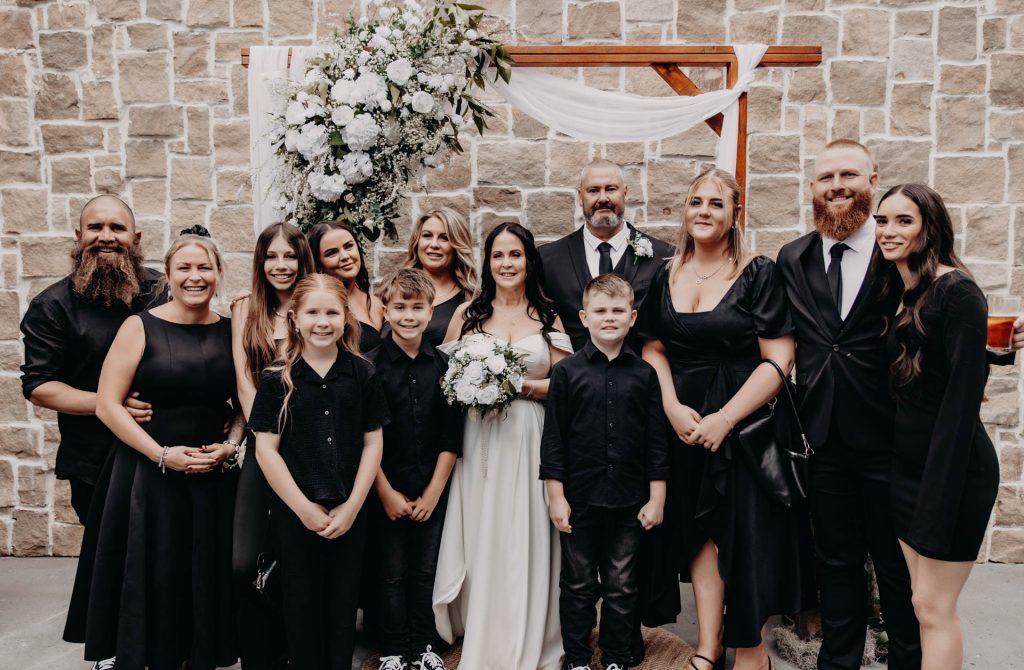 A group of people dressed in black and white stand together smiling in front of a floral wedding arch and a stone wall, with a bride and groom in the centre holding a bouquet.
