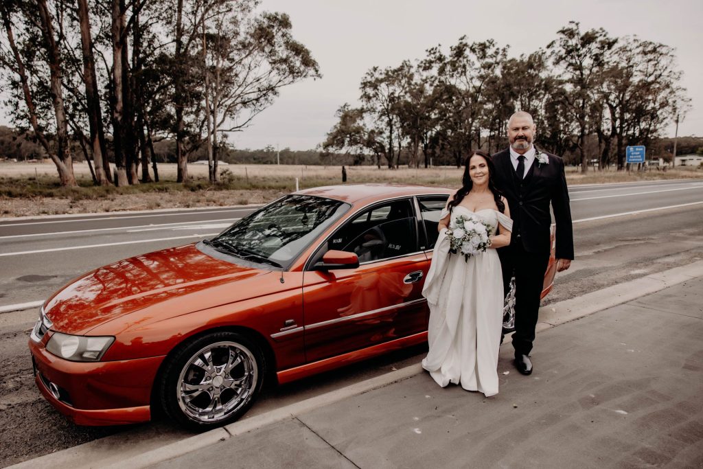 A bride and groom stand on a roadside next to a shiny red car. The bride wears a white dress and holds a bouquet, while the groom is in a black suit. Trees and a motorway are visible in the background.