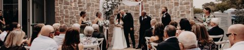 Renae and Wade stand under a floral arch, holding hands during their outdoor wedding ceremony, surrounded by bridesmaids, groomsmen, and seated guests facing the happy couple.