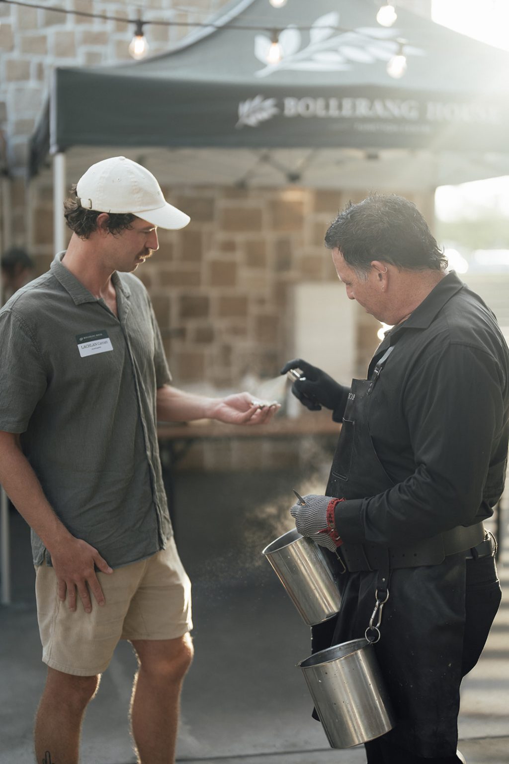 At a Bollerang House launch celebration, a man in a grey shirt and white cap receives a seasoning sprinkle on his palm from a man in black with gloves, holding two metal containers, at an outdoor event with marquee and lights behind them.