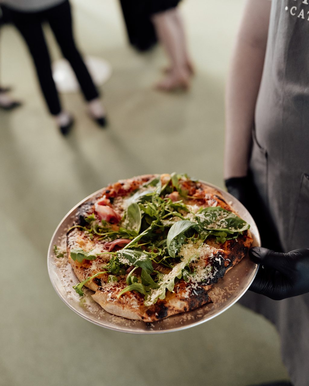 A person wearing a grey apron and black glove holds a plate with a rustic pizza topped with fresh greens and grated cheese at the Bollerang House launch. Blurred figures stand in the background on a light green floor.