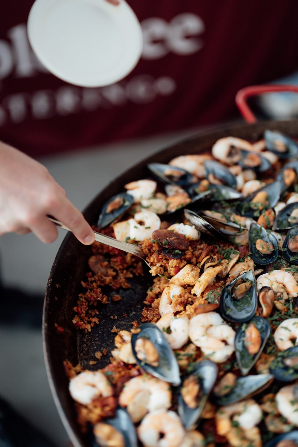 At Bollerang House, a hand scoops a serving of seafood paella from a large pan filled with mussels, prawns, rice, and vegetables—a perfect dish for any celebration or special launch.