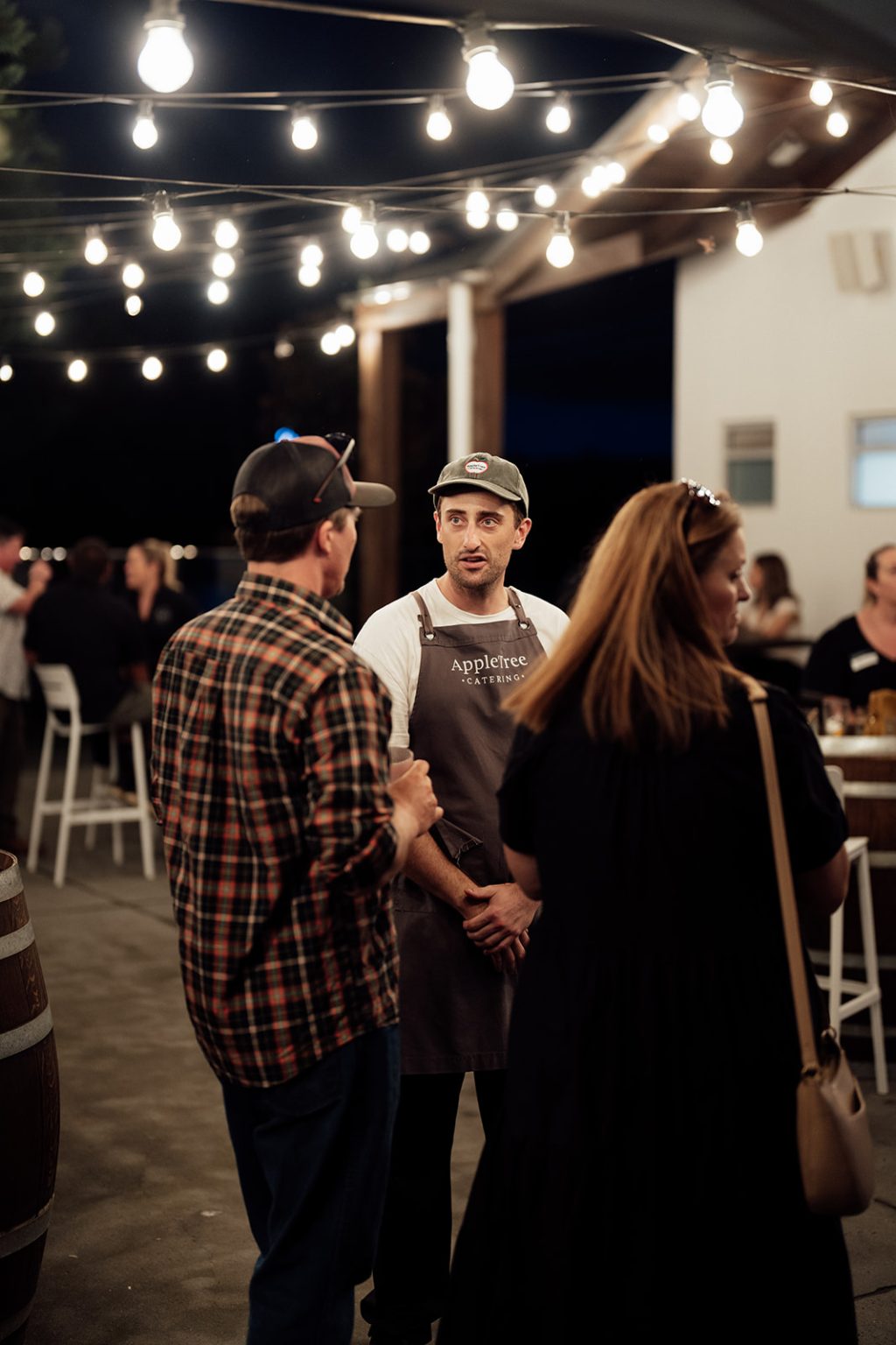 A man wearing an apron and cap stands under fairy lights, talking with two people at an outdoor evening gathering, celebrating the launch of Bollerang House. Other guests sit and mingle in the softly lit background.