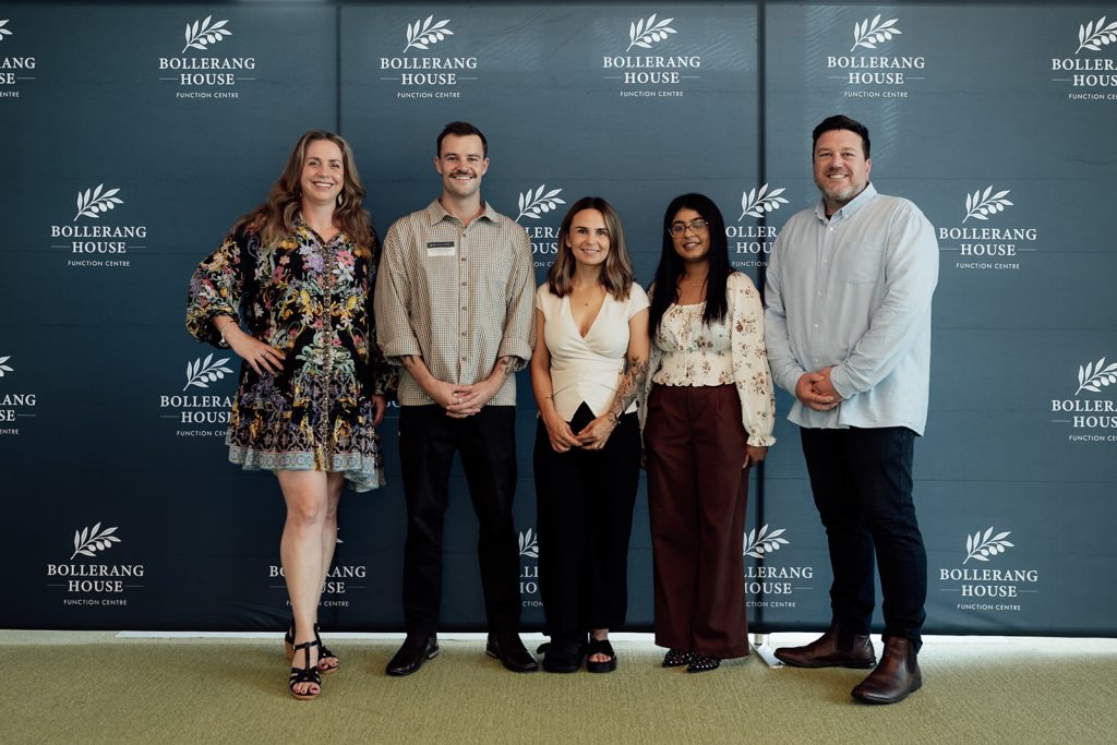 Five people stand smiling in front of a blue backdrop with “Bollerang House Function Centre” and a leaf logo. Dressed in business casual attire, they gather to celebrate the launch of Bollerang House.