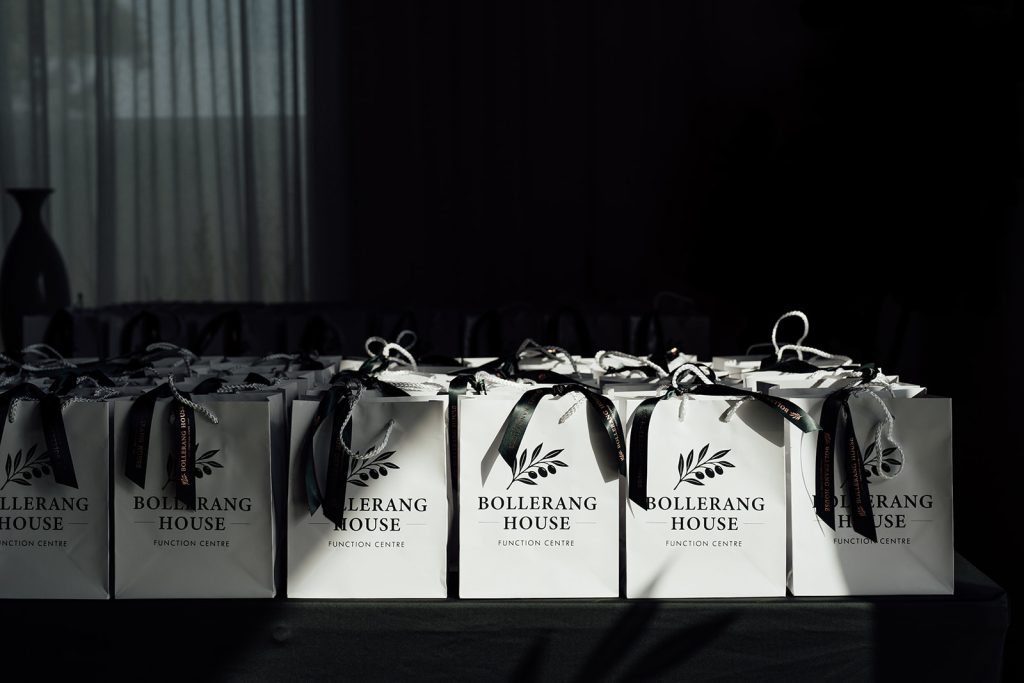 A row of white gift bags with green ribbons, each labelled "Bollerang House Function Centre," arranged neatly on a table in a dimly lit room, ready for the launch at Bollerang House.