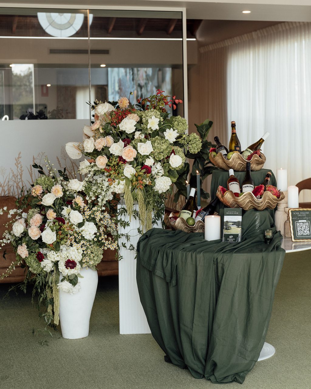 A decorated display at Bollerang House features large floral arrangements in white, peach, and green beside a table draped in dark green cloth, celebrating the launch with bottles of wine nestled in hessian, candles, and a small QR code stand.